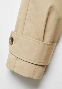Manica di un cappotto trench beige con cinturino regolabile e dettaglio con bottoni marroni scuri, che mostra cuciture ordinate e la trama del tessuto.