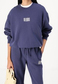Marineblauwe sweatshirt met ribgebreide boorden, met een witte geborduurde "BILLABONG" logo op de borst. Gepaard met bijpassende joggers en een beige tas.