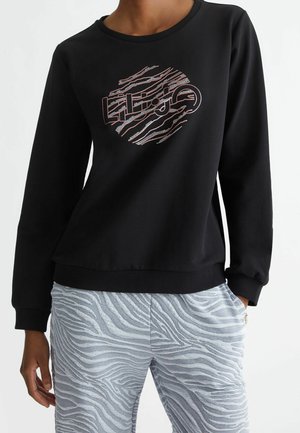 Personne portant un sweat-shirt noir avec un logo abstrait orange et blanc, un pantalon gris clair à motif zèbre, la main dans la poche.