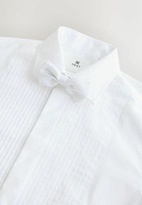 Valge särg, millel on volditud esiosa, volditud krae ja sobiv valge libliktie, millel on tekstuuriline täppmuster. Brändi etikett on nähtav.