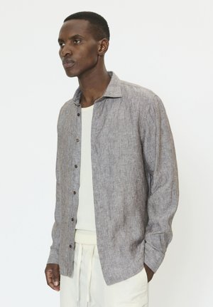 Mees, kes kannab heledat halli nööbitavat särk-jopet valge t-särgi ja valgete kootud pükstega, seisab ühevärvilise valge tausta ees.