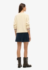 Suéter beige de punto con puños a rayas, combinado con una falda plisada azul oscuro y botas de cuero marrón hasta la rodilla con hebillas.