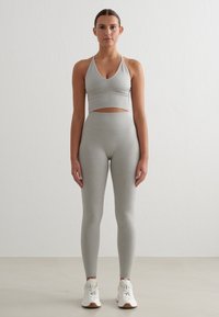 Šedá sportovní podprsenka s výstřihem do V a průstřihy, sladěná s šedými legínami s vysokým pasem a bílými teniskami. Hladký, elastický materiál.