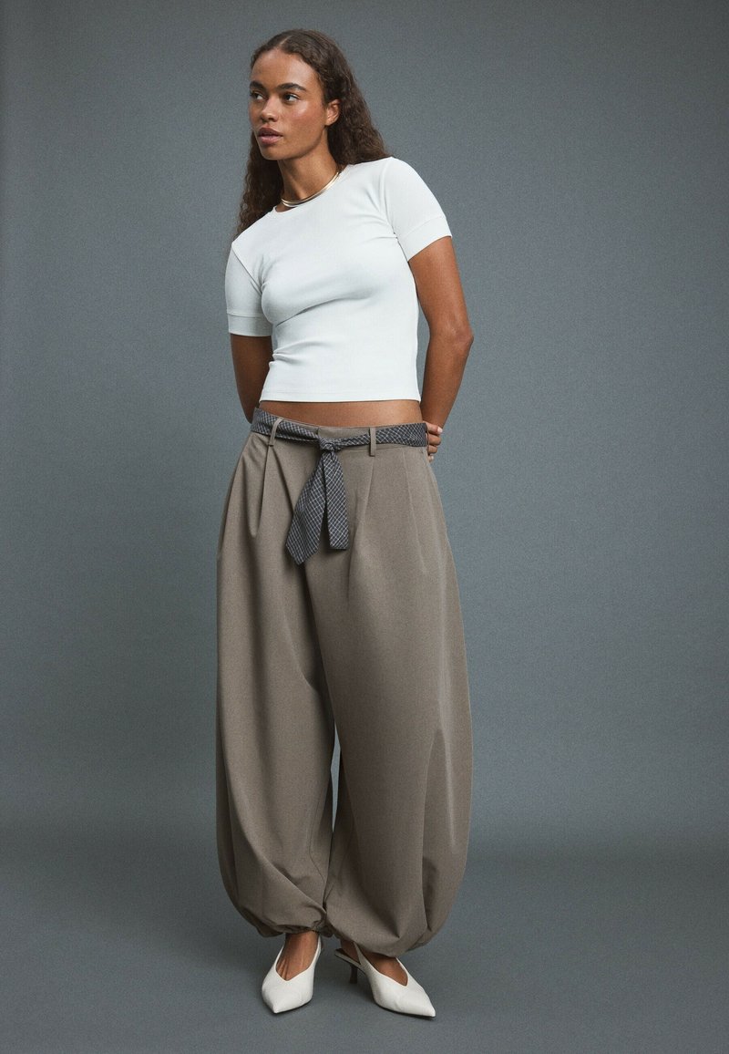 Femme aux longs cheveux bouclés porte un haut blanc cintré à manches courtes, un large pantalon beige avec ceinture nouée et des talons blancs pointus sur fond gris.