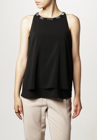 Svart ärmlös topp med lagerdesign, prydd med en krage av svarta och silverfärgade pärlor, tillverkad av ett slätt tyg. Bärs med ljusa byxor.