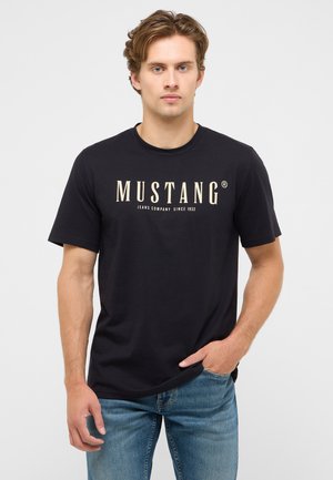 Junger Mann trägt ein schwarzes Mustang-T-Shirt mit dem Text "Jeans Company Since 1932", kombiniert mit blauen Jeans, steht vor einem einfarbigen Hintergrund.
