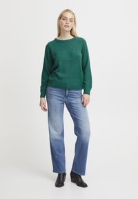 Maglione verde con texture, colletto rotondo, vestibilità comoda e polsini a coste, abbinato a jeans a gamba dritta di colore blu chiaro e stivali neri con tacco.
