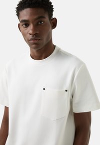 Biała koszulka z krótkim rękawem i okrągłym dekoltem, z kieszenią na piersi i ciemnymi, kontrastującymi guzikami. Gładka faktura, prosty design.