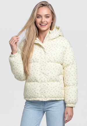 Młoda kobieta z długimi blond włosami, ubrana w jasnoyellową pikowaną kurtkę w kwiaty i jasnoniebieskie dżinsy, uśmiecha się i trzyma włosy.