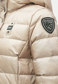 Giacca a vento beige con design trapuntato, texture lucida e una toppa nera con il marchio sulla spalla sinistra. Chiusura anteriore a snap.