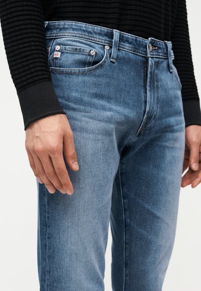 Άνδρας που φοράει μεσαίου τόνου μπλε τζιν και μαύρο μακρυμάνικο πουκάμισο με υφή, με τα χέρια χαλαρά στο πλάι, μπροστά σε απλό φόντο.