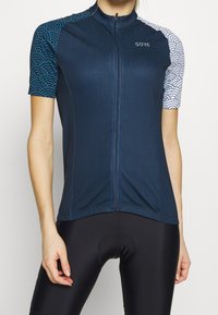 Kurzärmliges Radtrikot in Dunkelblau mit strukturiertem Körper; gemusterte Ärmel in Hellblau und Weiß. Voller Reißverschluss vorne. Leichtes Material.