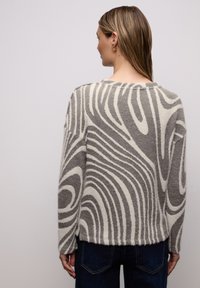 Pull en tricot gris et blanc à motif avec une coupe ample, des manches oversize et un ourlet côtelé. Le design présente des lignes ondulées et une texture douce.