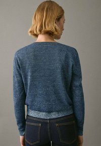 Kvinna med kort blont hår som bär en spräcklig blå långärmad tröja och mörka jeans, står vänd mot en enfärgad grå vägg.