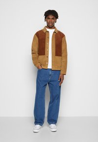 Nuori mies seisoo päällä kahden sävyinen ruskea antura takki, valkoinen paita, väljät siniset farkut ja valkoiset tennarit yksinkertaista harmaata taustaa vasten.