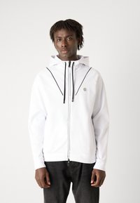 Haut à capuche blanc avec une fermeture éclair à l'avant, accent noir et logo sur la poitrine. Présente une capuche avec cordon de serrage et une coupe décontractée. Pantalon noir porté en dessous.
