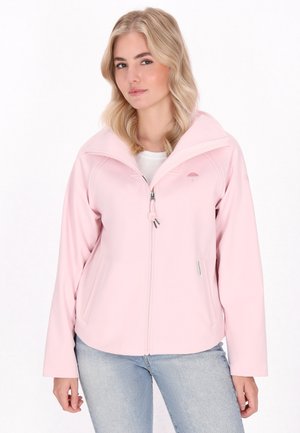 Ung kvinde med blonde hår iført en lyserød zip-up jakke, står foran en hvid baggrund og kigger direkte ind i kameraet.