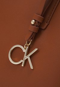 Ruskean keinonahkainen laukku, jossa on kullanvärinen "CK"-embleemiriipus kiinnitettynä vastaavan värisellä hihnalla. Laukun pinta on teksturoitu ja siinä on hienovaraiset pistotyyppiset yksityiskohdat.