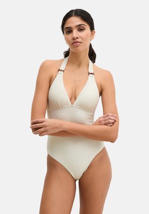 Donna con capelli scuri raccolti in una coda bassa, che indossa un costume intero avorio testurizzato con collo a fascia, braccia incrociate, su uno sfondo chiaro e uniforme.