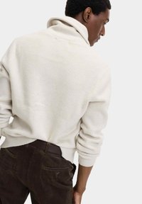 Weißer strukturierter Strickpullover mit hohem Kragen, getragen über dunkelbraunen Cordhosen. Zeigt die Rückansicht und ein entspanntes Schnittdesign.