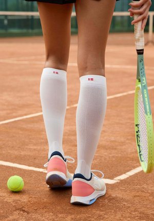 Valkoiset polvisukat punaisilla yksityiskohdilla, yhdistettynä vaaleanpunaisiin ja laivastonsinisiin tennareihin. Vihreä tennismaila ja keltainen tennispallo savikentällä.