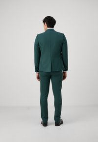 Costume vert au design ajusté, comprenant un blazer structuré et un pantalon taillé. Texture lisse, accents minimalistes, et sans motifs visibles.
