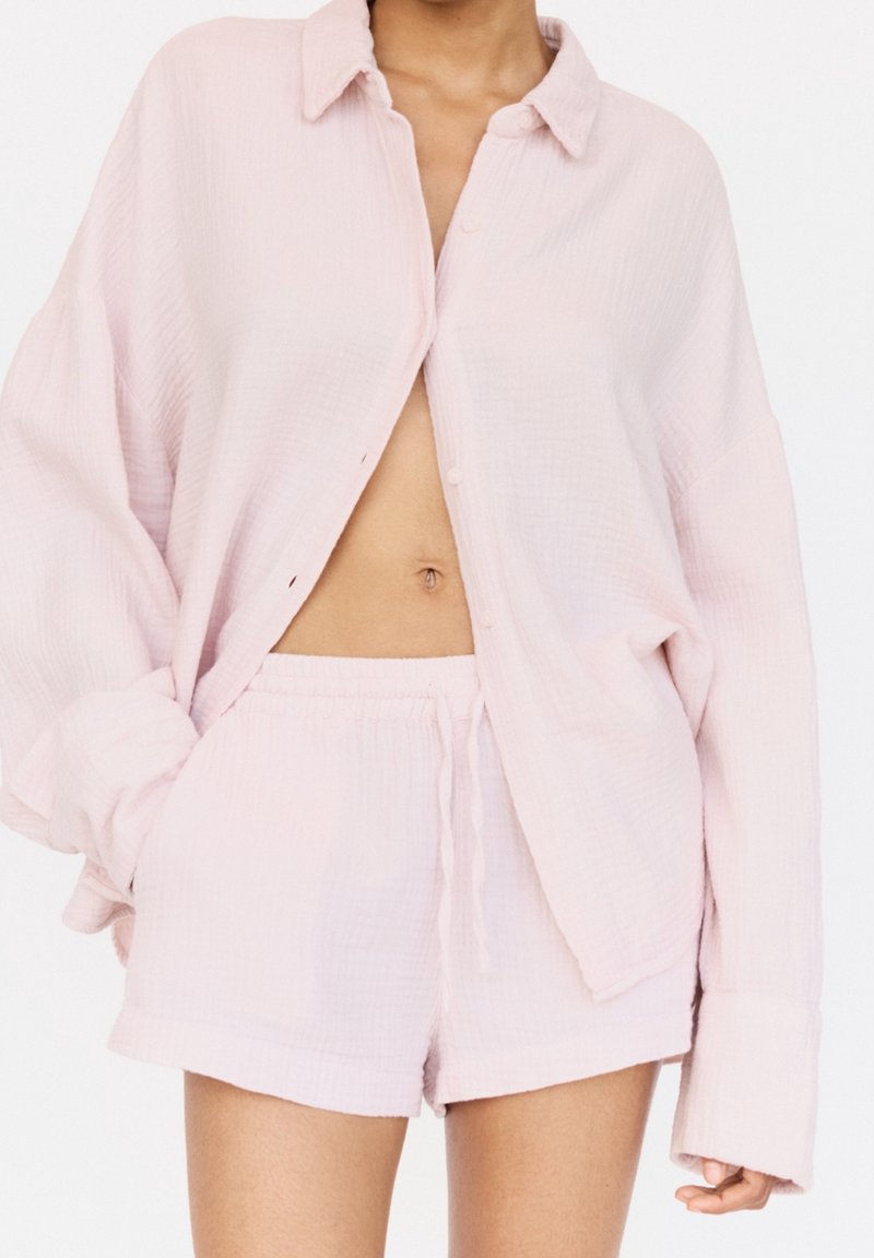 Personne portant une chemise ample rose clair boutonnée et un short assorti à cordon, montrant le ventre avec les mains détendues sur les côtés.