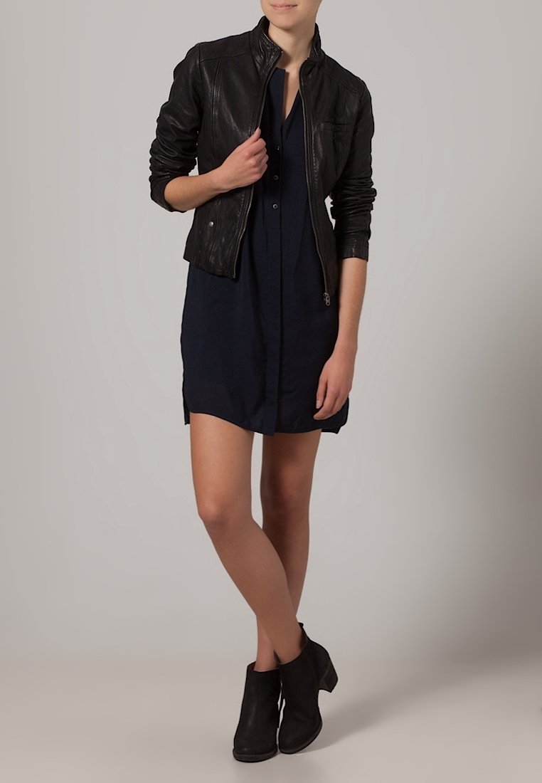 Czarna skórzana kurtka z zamkiem z przodu, noszona na ciemnoniebieskiej sukience koszulowej, zestawiona z czarnymi botkami za kostkę. Gładka faktura, dopasowany fason.
