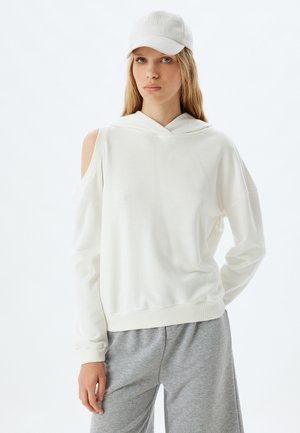 Sudadera blanca con capucha y diseño de hombros descubiertos, hecha de una tela suave. Combinada con pantalones de chándal grises para un look casual.