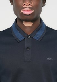 Pólo escuro com colarinho estruturado, apresentando detalhes em azul, dois botões e um discreto logótipo "BOSS" no peito. Textura de tecido suave.