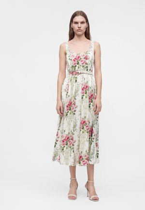 Femme debout dans une robe blanche sans manches à imprimé floral rose et vert, portant des sandales beiges à talons hauts sur un fond blanc uni.