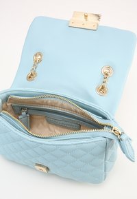 Ljusblått quiltat läderhandtag med guldhardware, med magnetlås och inre dragkedjefack. Klätt med beige tyg.