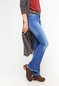 Jeans de campana azules, de lavado claro, combinados con una blusa color óxido, un cárdigan gris y zuecos de madera marrones con un acabado suave.