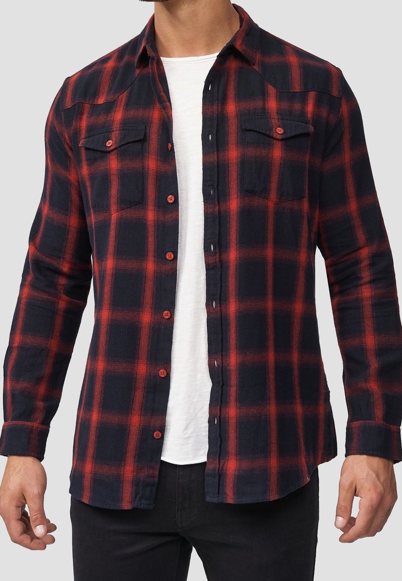 Mies, który ma na sobie rozpiętą czarną i czerwoną koszulę w kratę na białym T-shircie oraz czarne spodnie, stoi na jednolitym tle.