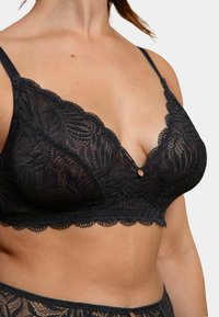 Bralette en dentelle noire avec un design à bords festonnés, décolleté en V profond et détail décoratif au centre. Tissu léger avec un motif floral complexe.