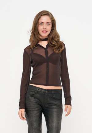 Jeune femme aux longs cheveux porte un haut noir transparent boutonné et un pantalon noir, souriant légèrement sur un fond clair uni.