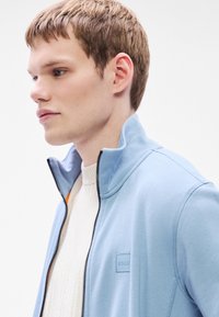 Ανοιχτό μπλε τζάκετ με φερμουάρ και γιακά τύπου σταντ, με διακριτικό λογότυπο, φορεμένο πάνω από κρεμ ριμπ πλεκτή μπλούζα. Ομαλή υφή υφάσματος.