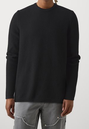 Homme portant un pull noir texturé à manches longues et un jean gris clair déchiré avec des détails de couture apparents.
