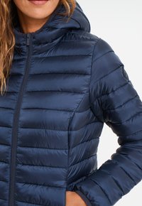 Chaqueta acolchada azul marino con capucha, que presenta costuras horizontales, un cierre frontal y una textura suave y brillante. Acabada con un logo en la manga.