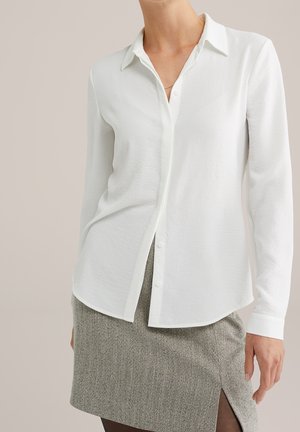 Kvinde iført en hvid skjorte med lange ærmer og knapper, stoppet ned i en grå fiskeben-nederdel med en frontslids.