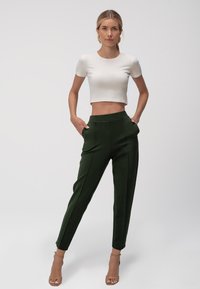 T-shirt bianco corto abbinato a pantaloni alti di un verde scuro, dotati di tasche laterali, tessuto liscio e gambe affusolate.