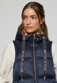 Gilet doudoune bleu marine avec un col haut et des accents dorés rose, portant le logo "POLO CLUB" et une fermeture éclair sur le devant. Design matelassé, texture lisse.