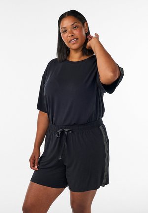 Femme à la peau mate et aux cheveux noirs raides portant un t-shirt noir à manches courtes et un short noir avec un cordon sur fond blanc.