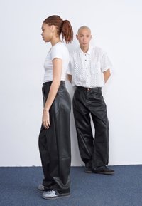 Černé kožené široké kalhoty s hladkou texturou, které nosí modelka, doplněné úzkým bílým tričkem a bílou perforovanou košili.