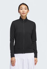 Fekete cipzáras dzseki texturált mintával, magas nyakrésszel és testhezálló kialakítással. Bordás mandzsettákkal és aljjal. Fehér szoknya fölött viselve.