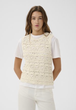 Mladá žena nosiaca krémový háčkovaný tielkový top cez biele tričko a svetlé nohavice, stojaca proti jednému pozadiu.