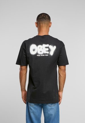 Svart bomull T-skjorte med korte ermer, med hvit spraymaling-stil "OBEY"-tekst på baksiden og ekstra tekst under.