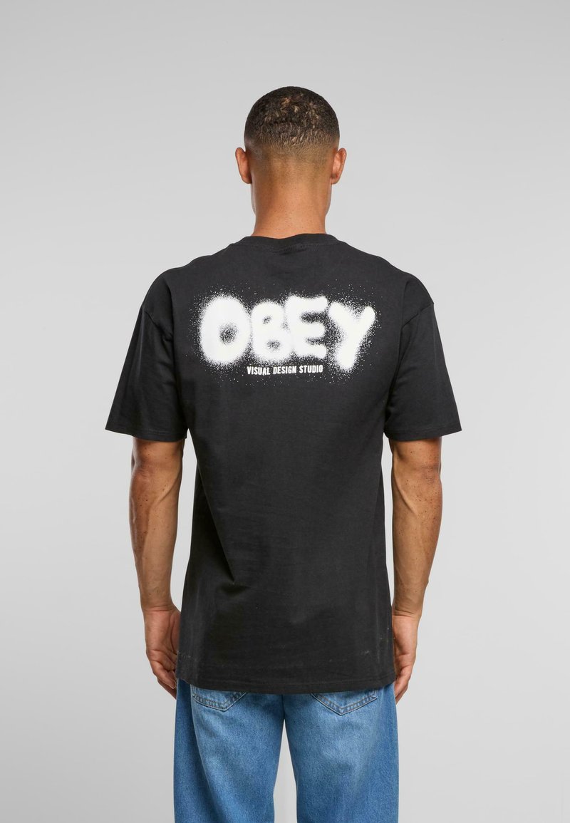 Melns kokvilnas T-krekls ar īsām piedurknēm, aizmugurē ar baltu izsmidzināšanas stila "OBEY" tekstu un papildu tekstu zem tā.