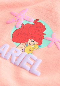 Ariel con pelo rojo abrazando un pez amarillo, decoraciones de estrellas de mar rosas y letras blancas que forman la palabra Ariel sobre tela rosa.