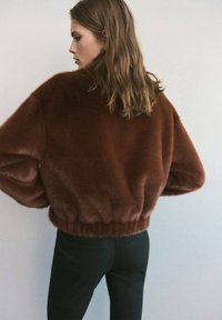 Giacca in pelliccia marrone con un design cropped, caratterizzata da una texture morbida, maniche ampie e orlo arricciato. Abbinata a pantaloni neri.